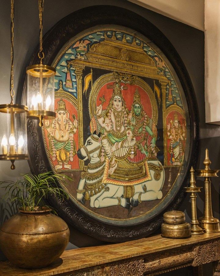 Traditional Kerala mural painting framed in a large circular wooden frame, depicting deities in vibrant colors, surrounded by brass lamps and décor on a carved wooden console — blending sacred art with timeless craftsmanship.