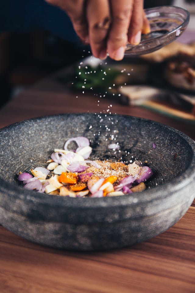 Close-up of a hand sprinkling salt into a stone mortar filled with sliced onions, garlic, and turmeric, preparing fresh ingredients for cooking.